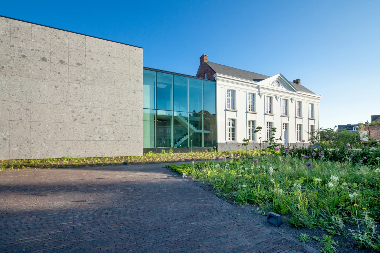 The office building of project G01: a renovated white presbytery with a glass link and a natural stone extension, with a flower meadow in front