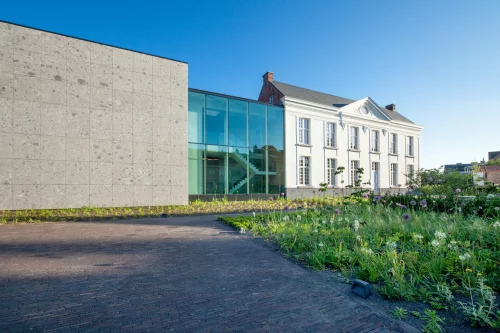 Het kantoorgebouw van project G01: een gerenoveerde witte pastorij met een glazen tussenstuk en een uitbreiding in natuursteen, met bloemenweide ervoor