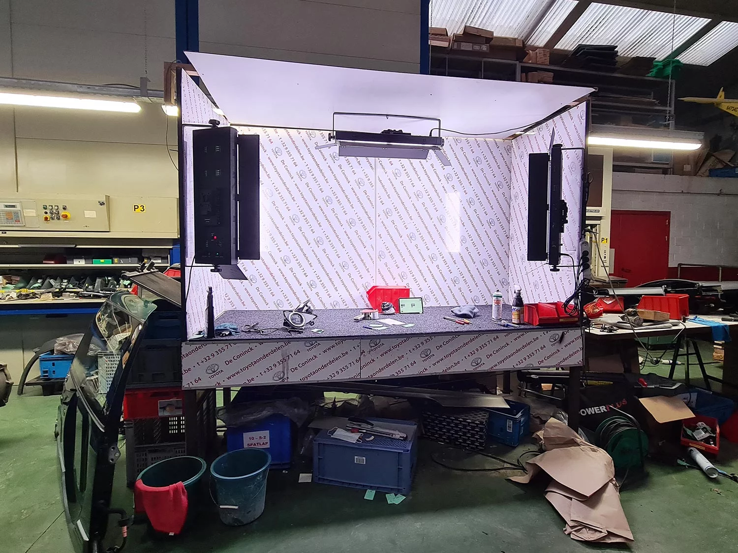 The photo studio inside the Toyota Parts De Coninck warehouse: a table with lamps and a softbox against self-printed wallpaper covered in Toyota logos